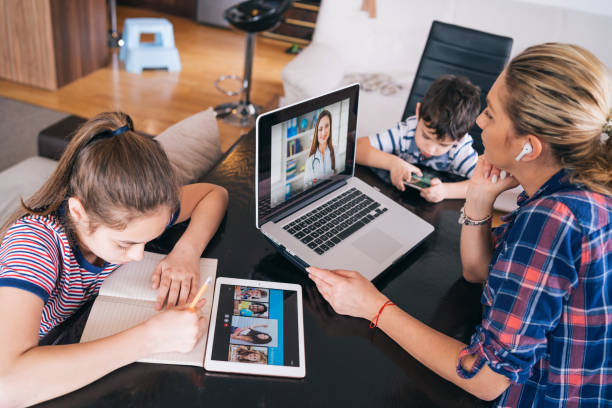 두 아이가 홈 스쿨링과 원격 학습을 머물면서 노트북에서 의사와 이야기하려고하는 어머니 - 멀리 떨어진 뉴스 사진 이미지