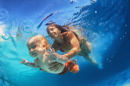 In blue pool young mother swimming with happy baby son - dive underwater with cheerful boy. Healthy family lifestyle and children water sports activity with parents during summer vacation with child