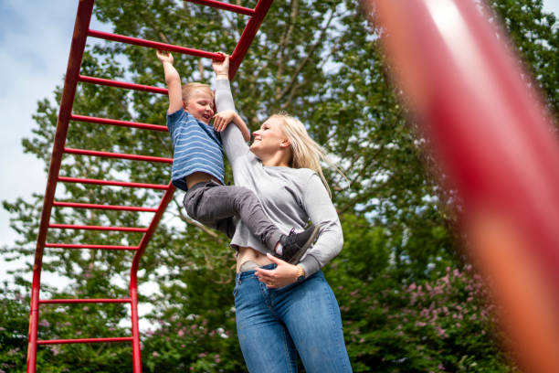 Mother with stoma bag assisting boy on monkey bars against trees stock photo