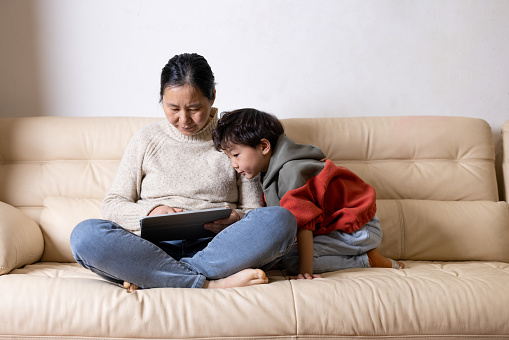 Asian mother sitting on sofa and working with tablet. His child sat beside him and watched. The mother is wearing a yellow sweater and the son is wearing a red sweater. During the pandemic, people try to live and work from home.