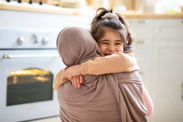 Mother's Day. Cheerful Little Girl Hugging Tight Her Muslim Mom In Hijab Mother's Day Concept. Cheerful Little Girl Hugging Tight Her Muslim Mom In Hijab, Islamic Woman Embracing Smiling Female Child, Happy Family Bonding Together In Kitchen At Home, Copy Space hot arabic girl stock pictures, royalty-free photos & images
