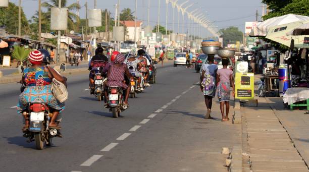 Motorbike taxis on the highway of Avepozo, Togo, West Africa. Avepozo, Togo - November 30, 2019: Motorbike taxis on the highway of Avepozo. The motorbike driver wear helmets, but the customers have no helmets. Vendors carry their goods on their head and walk at the highway. Location: Avepozo, Togo, West Africa. togo stock pictures, royalty-free photos & images