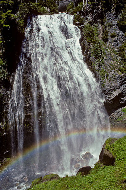 Rainbow Formed by a Waterfall Mount Rainier's numerous waterfalls are best viewed in early summer as melting snow feeds the streams, and again in autumn as the rains fill the streambeds. During late summer, only the major waterfalls will be flowing. Only a small number of the many waterfalls near Mount Rainier have been named. Whether the falls have names or not, they are a refreshing sight to both the eye and spirit. Narada Falls with its colorful rainbow were photographed near Paradise in Mount Rainier National Park, Washington State, USA. jeff goulden rainbow stock pictures, royalty-free photos & images