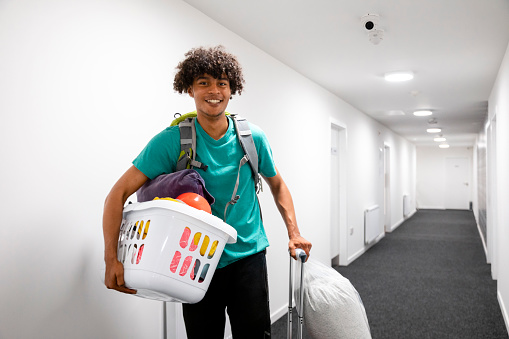 A medium close-up of a young male student with a smile on his face as he makes his way to his new room in a student accommodation building in Sunderland in the North East of England. He is carrying belongings with him and looking into the camera with a smile on his face.