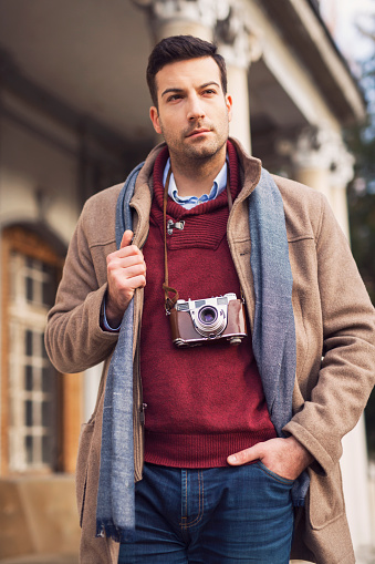 Young, handsome man carying his retro camera around the neck and standing in front of the old, luxurious house