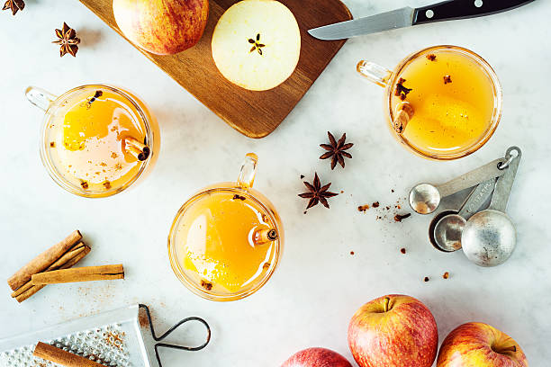 Mugs of Hot Spiced Mulled Apple Cider with Ingredients Three glass mugs of hot mulled apple cider heated with cinnamon sticks, star anise, and cloves on a marble countertop. Apples are on a wooden cutting board ready to be sliced and a grater is on the side for the spices. cider stock pictures, royalty-free photos & images