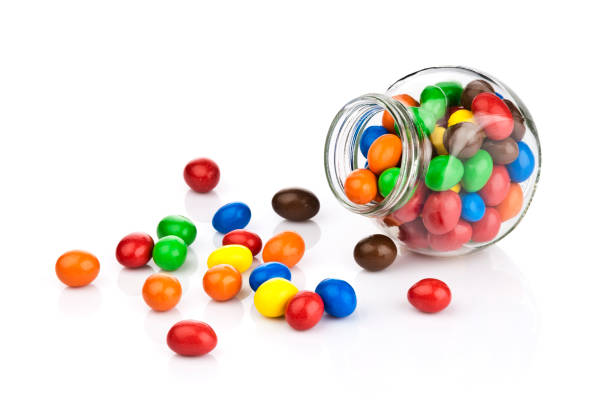 Multi colored chocolate covered candies on white background Open candy jar filled with multi colored chocolate covered peanut candies on white background. Some candies are spilled out of the jar directly on the background. DSRL studio photo taken with Canon EOS 5D Mk II and Canon EF 70-200mm f/2.8L IS II USM Telephoto Zoom Lens candy jar stock pictures, royalty-free photos & images