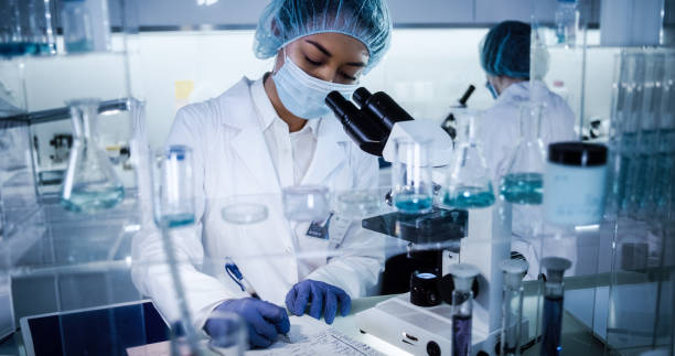 Multi ethnic, female team studying DNA mutations. Using microscope in protective workwear Scientists examines DNA models in modern Genetic Research Laboratory. Computer monitors with data in foreground biotechnology photos stock pictures, royalty-free photos & images