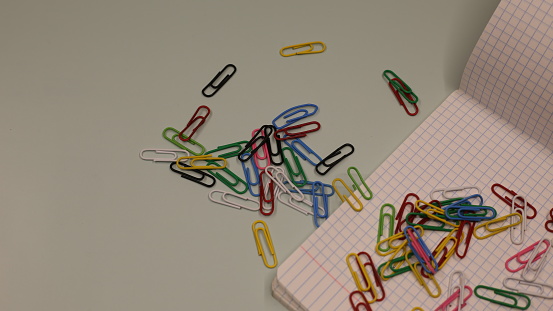 Multi-colored paper clips. Many metal paper clips are scattered on the table. Notebook with staples. Bright background with colored paper clips