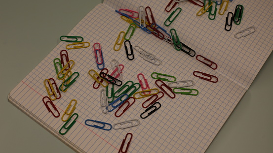 Multi-colored paper clips. Many metal paper clips are scattered on the table. Notebook with staples. Bright background with colored paper clips
