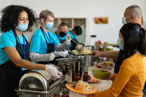 Kind helpers serving a healthy hot meal to disadvantaged families in time of poverty, wearing protective face masks, aprons, name tags and protective gloves for hygiene