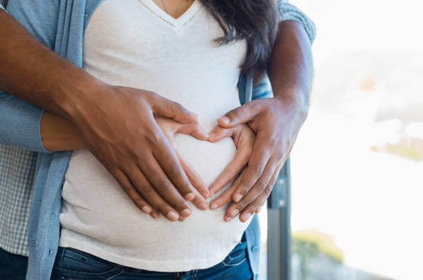 Multiethnic couple expecting a baby Pregnant woman and her african husband holding hand in heart shape on baby bump. Close up of multiethnic couple making heart shape on the tummy. Loving future couple expecting a baby. stomach photos stock pictures, royalty-free photos & images