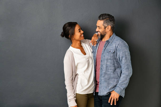 Multiethnic couple looking at each other with a smile Happy mature indian couple embracing and looking at each other against gray background. Mid adult multiethnic couple in love standing against grey wall. Middle eastern man fall in love of his beautiful hispanic girlfriend. mid adult couple stock pictures, royalty-free photos & images