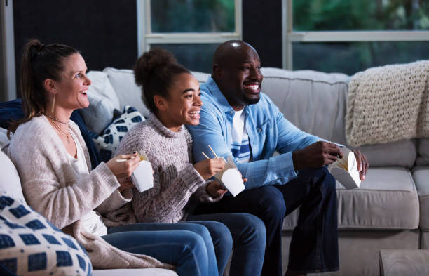 famille multiethnique sur le canapé en regardant la télévision, en mangeant des choses à emporter - african america man eating chinese food photos et images de collection