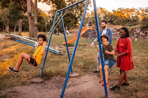 Multi-ethnic family of four playing on a swing in a public park on a sunny summer day.