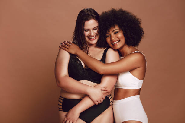 Multi-ethnic females in lingerie standing together Smiling young african female hugging her female friend on brown background. Multi-ethnic females in lingerie standing together and smiling. body positive stock pictures, royalty-free photos & images