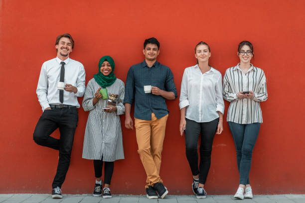 Multiethnic group of casual businesspeople using smartphone during a coffee break from work in front of the red wall outside. stock photo