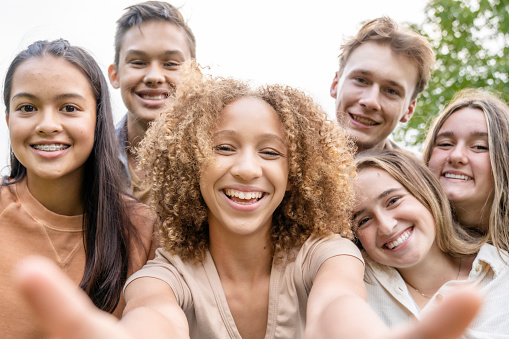 A group of ethnically diverse high school students stand closely together posing for a photo.  They are each dressed casually in hues of peach and pink as they smile.  The focus is on a curly haired girl in front who has her arms outstretched as she holds her cell phone to take a selfie of the group.