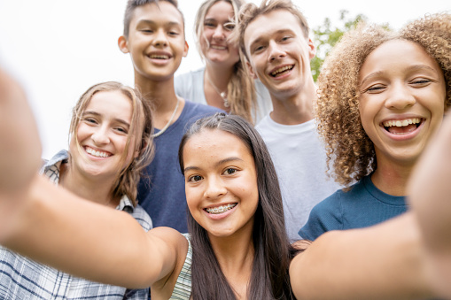 A group of ethnically diverse high school students stand closely together posing for a photo.  They are each dressed casually in hues of blue as they smile.  The focus is on a brunette girl in front who has her arms outstretched as she holds her cell phone to take a selfie of the group.
