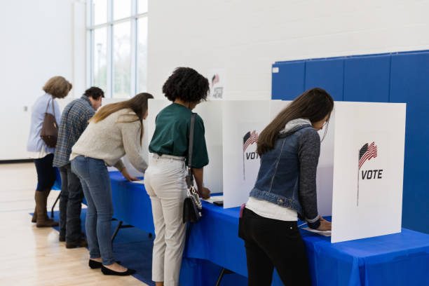 Multi-ethnic group votes at voting booths An unrecognizable mutli-ethnic group of voters stands to vote at the voting booths lined up against the wall of the gym. voting booth stock pictures, royalty-free photos & images