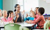 A multi-ethnic group of six preschool children with a mixed race African-American and Caucasian teacher, sitting around a table in a classroom. The teacher and some of her students have their hands raised, holding up fingers, learning how to count. The children are 4 years old.