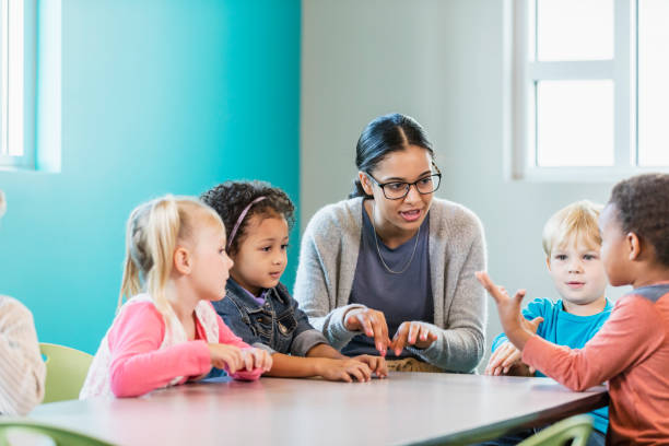 Multi-ethnic preschool teacher and students in classroom A multi-ethnic group of four preschool children with a mixed race African-American and Caucasian teacher, sitting around a table in a classroom.  The teacher, a young woman in her 20s, is looking at an African-American boy. The children are 4 years old. teach reading to kindergarteners stock pictures, royalty-free photos & images