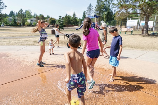 A group of mixed race children of varying ages play in the fountain at a public water park on a hot and sunny summer day. A young mother pushing a stroller and the kids' grandmother are in the background. A playground and homes surrounded by trees are in the distance.