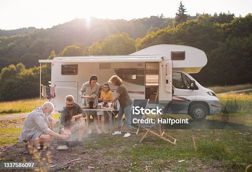 istock Multi-generation family sitting and eating outdoors by car, caravan holiday trip. 1337586097