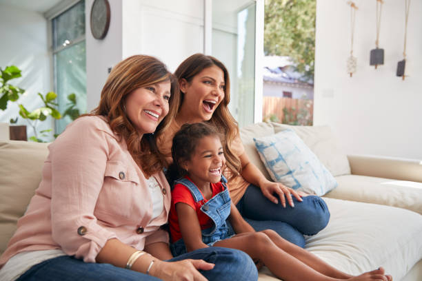 Multi-Generation Female Hispanic Family On Sofa At Home Watching TV Together Multi-Generation Female Hispanic Family On Sofa At Home Watching TV Together latin family stock pictures, royalty-free photos & images