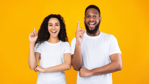 Multiracial Couple Pointing Fingers Up Having Idea Over Yellow Background stock photo