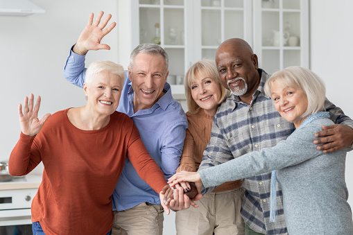 Multiracial group of happy senior friends men and women in casual outfits holding hands in top of each other and waving at camera, enjoying time together, kitchen interior. Elderly people lifestyle