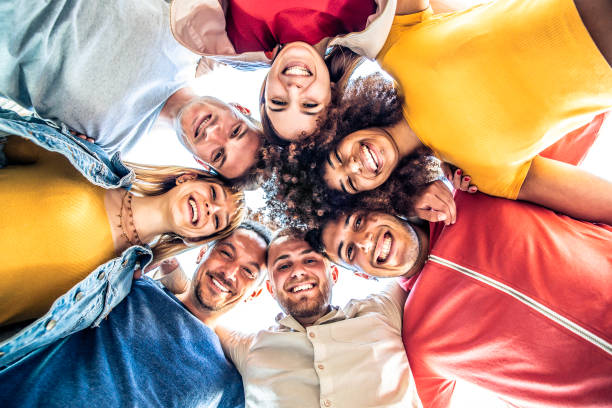 Multiracial group of young people standing in circle and smiling at camera - Happy diverse friends having fun hugging together - Low angle view stock photo