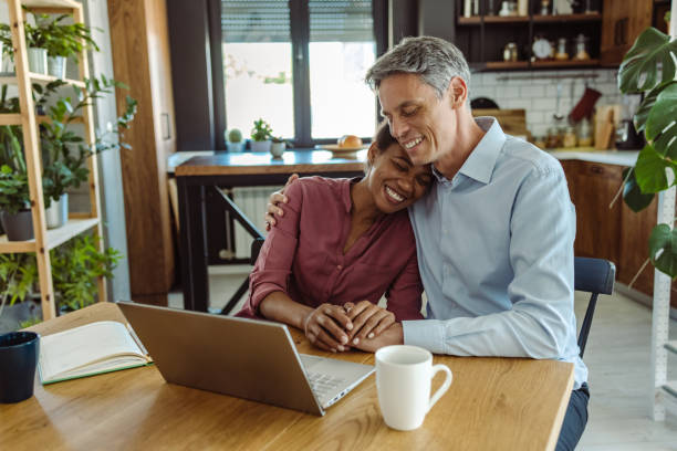 Multi-racial mature couple enjoying home and using laptop stock photo