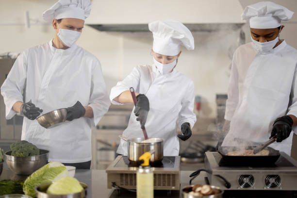 Multiracial team of cooks in uniform and face masks cooking meals for a restaurant in the kitchen stock photo
