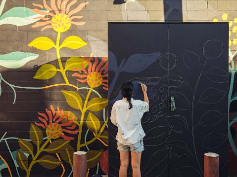 Young Asian woman, mural artist creating wall art at the urban setting.