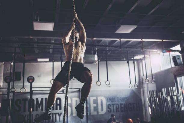 Muscular build athlete climbing up the rope in a gym. Young athletic man exercising strength on a rope in a gym. bodybuilder stock pictures, royalty-free photos & images