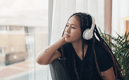 Shot of a young woman using headphones and looking thoughtfully out of a window at home