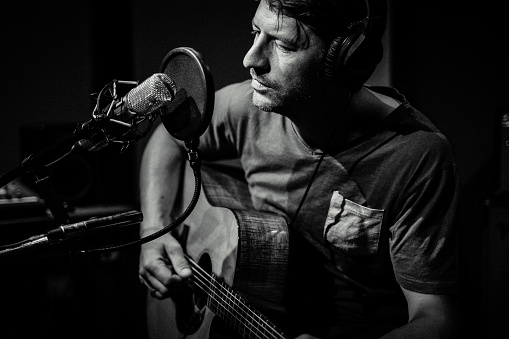 A Musician looks down at the microphone with his hands resting on his guitar. He is wearing headphones. Moody black and white image.