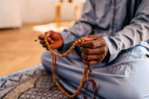 Young African-American Muslim man wearing kaftan holds prayer beads while praying at home.