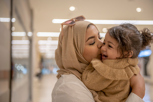 A young Muslim mother holds her daughter on her hip while leaning in to kiss her cheek.  They are both dressed in neutral colors and the mother is wearing a hijab.