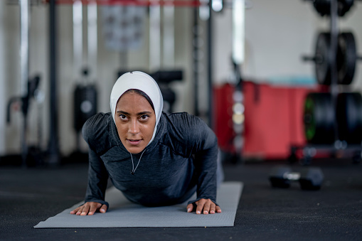 A young woman of Middle Eastern decent lays out on a yoga mat in a fitness center as she works out by herself.  She is dressed comfortably in athletic wear and focused on her push ups and breathing.