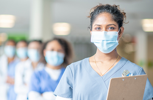 A female doctor of Indian descent is wearing grey medical scrubs and a face mask stands in front of her colleagues at the hospital. She is holding a clipboard and appears to be looking serious.