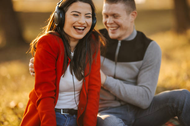 My greatest pleasure is to make her laugh Cheerful young heterosexual couple sitting and laughing on picnic blanket in nature at sunset and having fun while listening good music on headphones tickling beautiful women pictures stock pictures, royalty-free photos & images