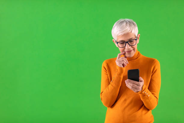 My kids sends me pictures everyday Senior woman wearing eyeglasses, working while using mobile phone against green background. smart phone green background stock pictures, royalty-free photos & images