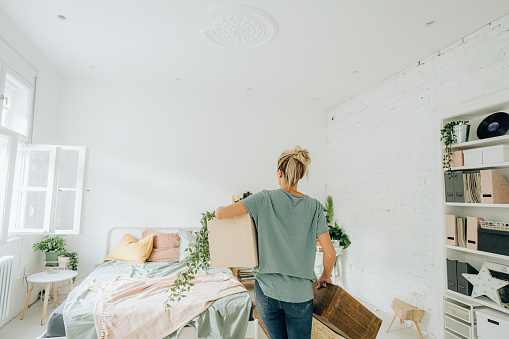 Photo of a young woman arranging the bedroom of her new apartment; carrying boxes and containers and planning her living space.