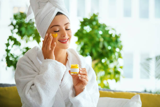 My skin is already feeling better. Young woman in bathrobe and wrapped in towel applying face cream. She's holding beauty products, smiling with her eyes closed. applying face cream stock pictures, royalty-free photos & images