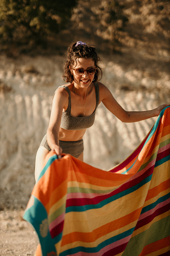 Beautiful smiling female in a bikini, spreading a towel on the beach, ready to enjoy in Sun by the lake/sea. Summer holiday and vacation concept