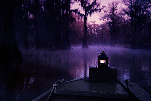 A boat with a lit lantern glides through an eerie blad cypress swamp at dawn with fog low on the water, Caddo Lake, on the border between Louisiana and Texas, USA