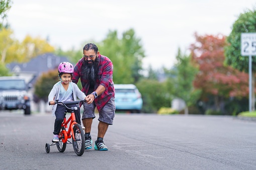 A supportive and loving dad patiently holds his six year old daughter steady while teaching her how to ride a bike in the street of their residential neighborhood. The young girl is wearing a helmet and her bike has training wheels.
