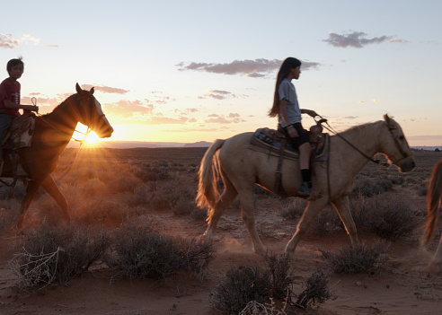 Native american navajo teenagers and children riding horses together in Monument Valley Arizona, Utah at dusk under a dramatic sunset in the late summer on the tribal park